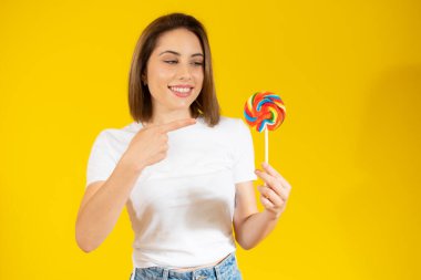 Elinde renkli lolipop şekeri tutan güzel genç bir kadın sarı arka planda eğleniyor. Tatlı yemek ve mutluluk konsepti.