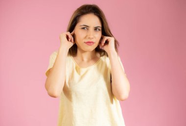 Gündelik tişört giyen genç ve güzel bir kadın izole edilmiş pembe arka planda dikiliyor kulakları rahatsız edici bir ifadeyle parmaklarıyla örtüyor. Sağır kavramı.
