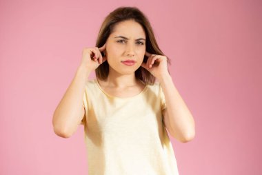 Gündelik tişört giyen genç ve güzel bir kadın izole edilmiş pembe arka planda dikiliyor kulakları rahatsız edici bir ifadeyle parmaklarıyla örtüyor. Sağır kavramı.