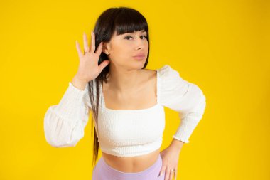 İzole edilmiş sarı arka planda genç esmer kadın elini kulağına koyarak bir şeyler dinliyor.