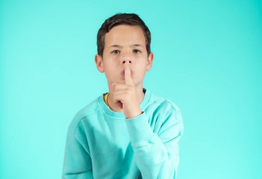 Kafkasyalı genç, yeşil bir stüdyo duvarında günlük kıyafetlerle ciddi bir şekilde poz vererek sessizliği işaret ediyor.