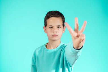 Beyaz küçük bir çocuk yeşil duvarın önünde duruyor ve üç numaralı parmaklarıyla işaret ediyor.