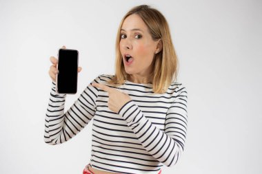 Beyaz arka planda tek başına dururken boş cep telefonu gösteren gülümseyen genç bir kadının portresi