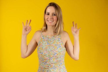 Sarı arka planda duran yaz elbisesi giyen genç ve güzel bir kadın gülümsüyor..