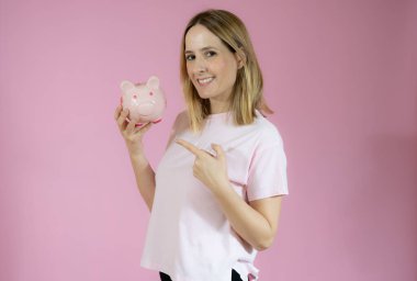 Güzel bir kadın pembe arka planda izole edilmiş kumbarayla gülümsüyor..