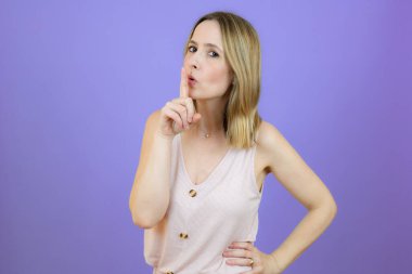 Portrait of beautiful woman standing over isolated purple background asking to be quiet with finger on lips. Silence and secret concept.
