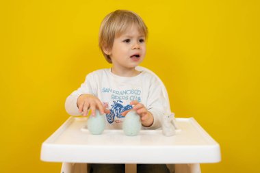 Güzel erkek bebek oturuyor ve sarı arka planda izole edilmiş iki paskalya yumurtası tutuyor..