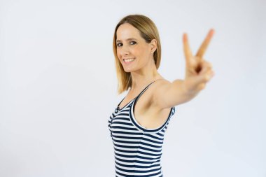 Çizgili elbiseli güzel, gülümseyen genç kadın beyaz arka planda izole edilmiş zafer işareti gösteriyor..