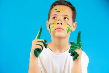 Cute boy with finger paint pointing up isolated over blue background.