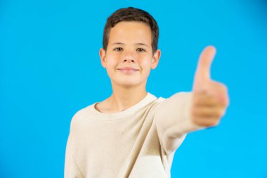 Smiling boy in casual sweater showing thumb up isolated over blue background.