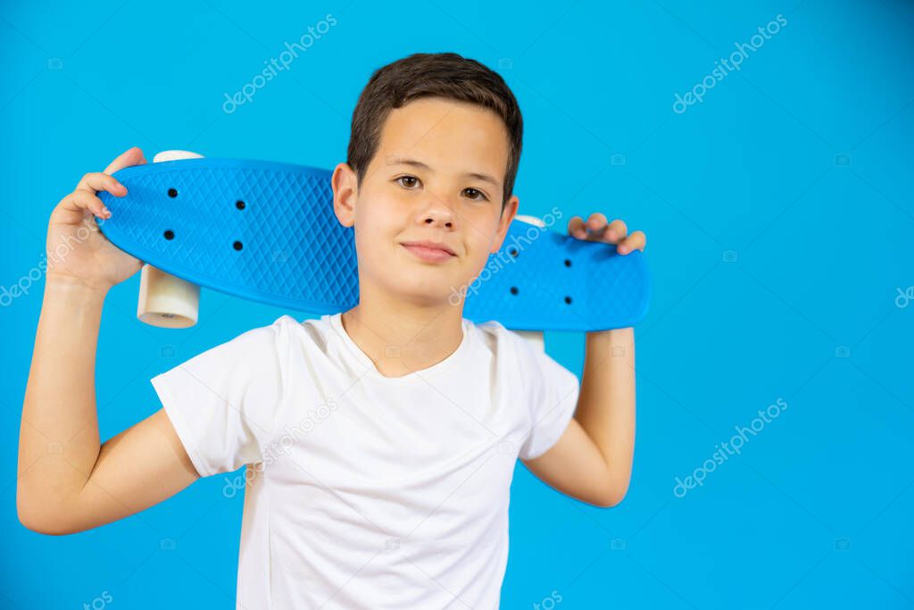 Young smiling boy holding skateboard isolated over blue background.