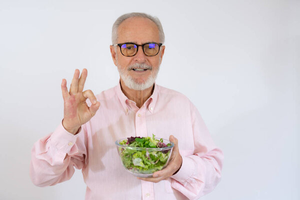 Веселый старший держит миску с салатом и глядя на камеру изолированы на белом фоне