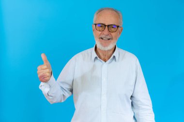 Gri saçlı son sınıf öğrencisi mavi arka planda dikilip gülümseyen ve başparmağıyla kameraya bakan..