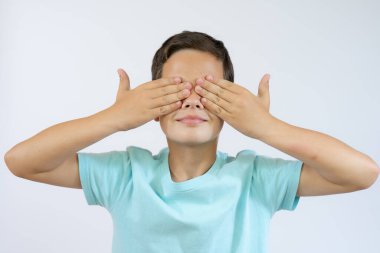 Mavi tişörtlü, elleri beyaz arka planda genç bir çocuk yüzünü kapatıyor..