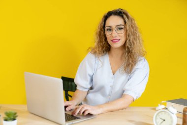 Bilgisayarda çalışan genç bir kadın sarı arka planda gülümsüyor..