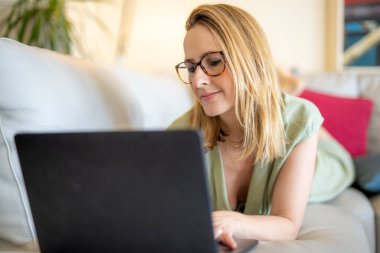 Dizüstü bilgisayarda çalışan ve evinde kanepede yatarken gülümseyen çekici genç bir kadın.