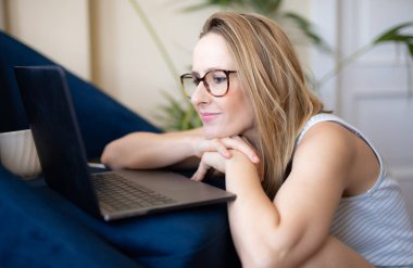 Dizüstü bilgisayarda çalışan ve evinde yerde otururken gülümseyen çekici genç bir kadın.