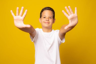 Beyaz tişörtlü, sarı duvara yaslanmış küçük şirin beyaz çocuk avuç içiyle el hareketi yapıyor.