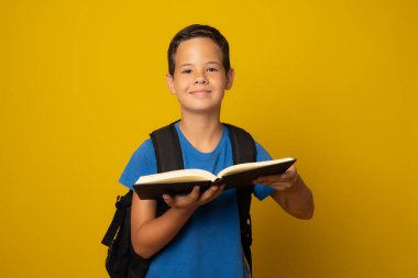 Sarı arka planda izole bir kitap tutan mutlu okul çocuğu..