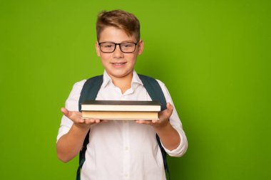 Sırt çantası ve yeşil arka planda saklanmış kitapları olan gülümseyen bir çocuk.