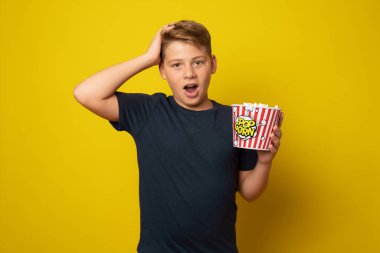 Büyük bir kutu patlamış mısır tutan neşeli çocuk sarı arka planda izole edilmiş kameraya bakıyor.