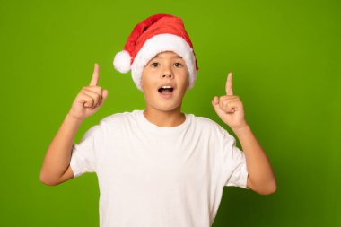 Kırmızı şapkalı ve beyaz tişörtlü, yeşil arka planı işaret eden şaşırmış bir çocuğun portresi. Noel Baba şapkalı bir çocuk..