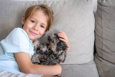 Portrait of a smiling caucasian child boy playing with his little puppy dog on the couch at home.