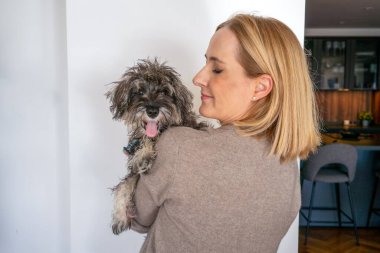 Evde gülümseyen sarışın bir kadının yavru köpeğine sarılışının yan görüntüsü..