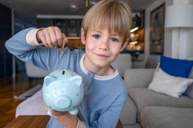Pijamalı, gülümseyen sarışın çocuk domuz kumbarasını alıp para biriktirmek için para koyuyor.