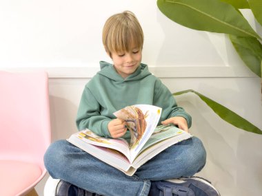 7 yaşında sarışın bir çocuk bir sandalyede oturur ve bir odada kitap okur. Eğitim kavramı.