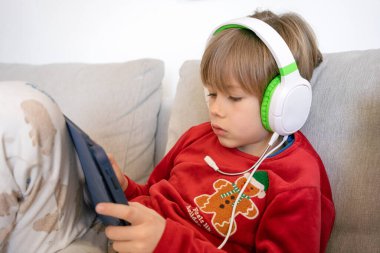 Çocuk, tablet kullanırken kulaklık takıyor, evdeki kanepede oturuyor. Teknoloji konsepti.
