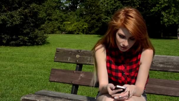 Young attractive woman sits on bech in the park and smiles - woman works on the smartphone (sms) - nature — Stock Video