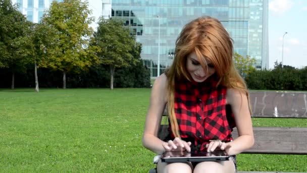Jovem mulher atraente trabalha no tablet (bate-papo) e sorri - senta-se no parque (natureza - grama e árvores) - edifício moderno — Vídeo de Stock