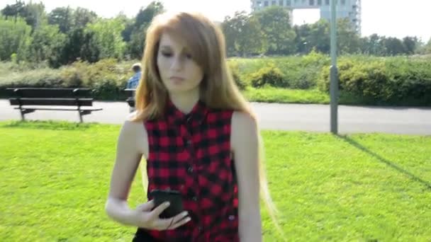 Young attractive woman goes in park and listens to music on smartphone - other people in the background with buildings and nature (tree and grass) — Stock Video