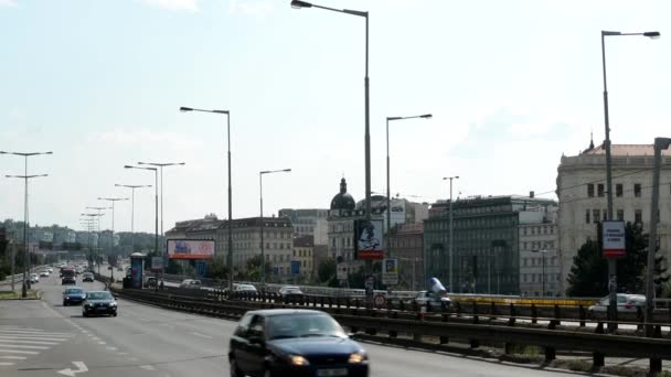 Urban street med förbipasserande bilar - byggnader i bakgrunden - natur (träd) - människor - molnig himmel (soliga) — Stockvideo