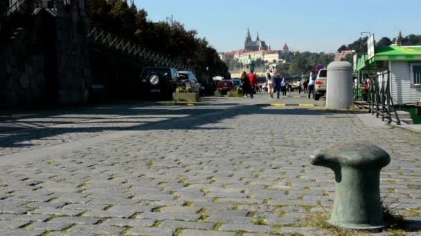 Wandelen mensen (jonge paar verliefd) en fietsers - waterfront - stad (gebouwen en bomen) op achtergrond - Kasteel van Praag - Hradcany - zonnige (blauwe hemel) — Stockvideo