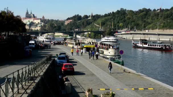 Boats on the river in quay (Vltava) - city (buildings) in background - Prague Castle (Hradcany) - sunny (blue sky) - cars and trees - walking people - waterfront — Stock Video