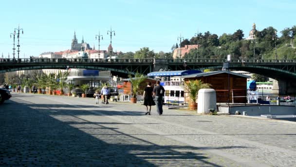 Boats on the river in quay (Vltava) - city (buildings) in background - Prague Castle (Hradcany) - sunny (blue sky) - cars and trees - walking people - waterfront - bridge — Stock Video