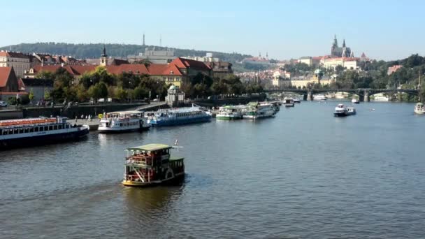 Barche sul fiume (Moldava) - città (edifici) sullo sfondo - Castello di Praga (Hradcany) - sole (cielo blu) - auto e alberi — Video Stock