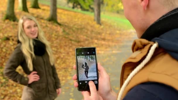 Pareja modelo joven enamorada - parque de otoño (naturaleza) - hombre fotografiando a mujer con smartphone - pareja feliz - primer plano — Vídeos de Stock