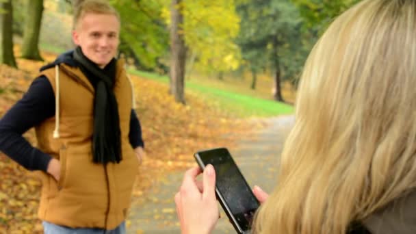 Pareja joven modelo enamorada - parque de otoño (naturaleza) - mujer fotografiando al hombre con smartphone - pareja feliz - primer plano — Vídeos de Stock