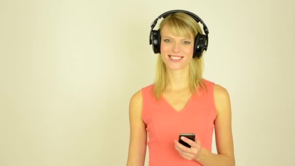 Fiatal, vonzó nő hallgat zenét a fejhallgatót a telefon - stúdió - nő néz ki, hogy a kamera — Stock videók