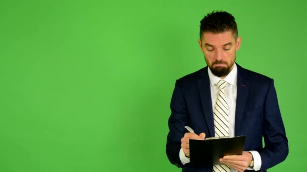 Biznes człowiek pisze do papieru i uśmiecha się do kamery - zielony ekran - studio — Wideo stockowe