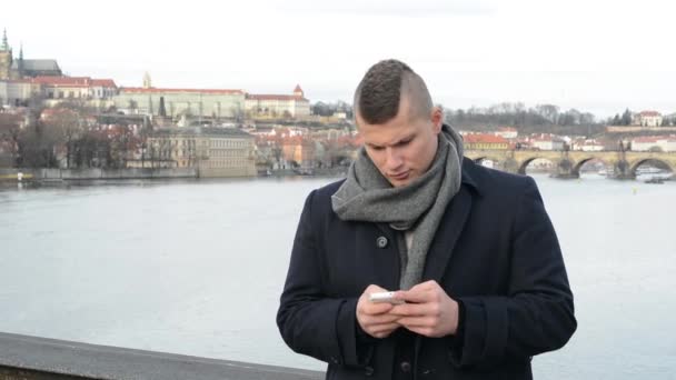 Homem trabalha no smartphone e sorri para a câmera na ponte - cidade (Praga) em segundo plano — Vídeo de Stock