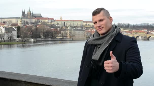 Young handsome man shows thumb on agreement and smiles to camera - city (Prague) in background — Stock Video