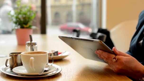 Mujer trabaja en la tableta en la cafetería - disparo en la mano - café y pastel en el fondo - calle urbana con coches en el fondo — Vídeos de Stock