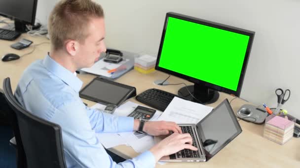 Joven hombre guapo trabaja en el ordenador portátil en la oficina - pantalla verde - computadora de escritorio y tableta — Vídeos de Stock