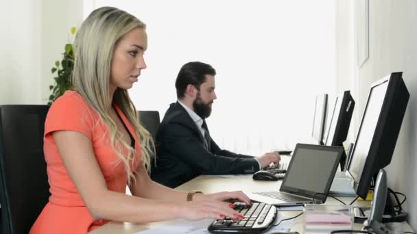 Junge attraktive Frau arbeitet im Büro am Computer und lächelt in die Kamera - Mann arbeitet am Desktop-Computer im Hintergrund — Stockvideo