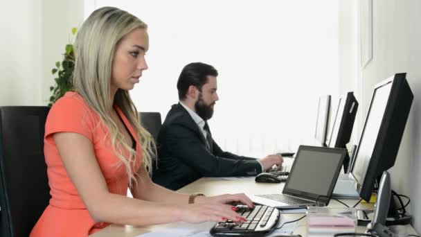 mladá atraktivní žena pracující na počítači v kanceláři a usmívá se do kamery - muž pracuje na stolním počítači v pozadí