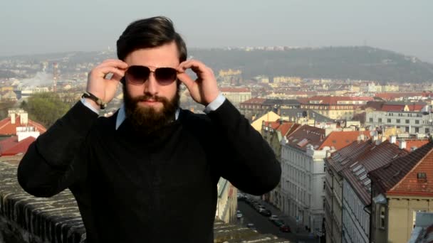 Young handsome man with full-beard (hipster) puts on his sunglasses and smiles - city in background — Stock Video